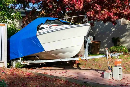 White boat covered with a blue tarp stored on a trailer in a residential driveway during fall.