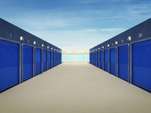 Symmetrical outdoor self-storage facility corridor with numbered blue roll-up doors on both sides under a clear blue sky.